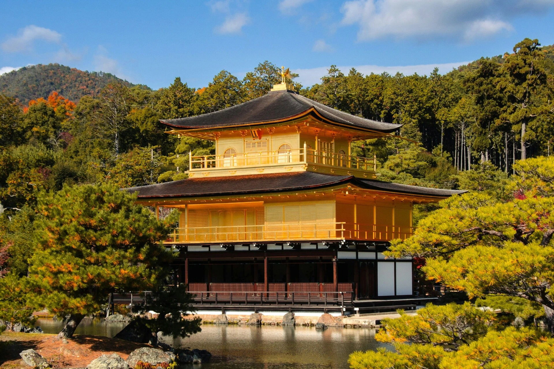 A golden pavilion surrounded by trees, with a pond in the foreground, under a partly cloudy blue sky.