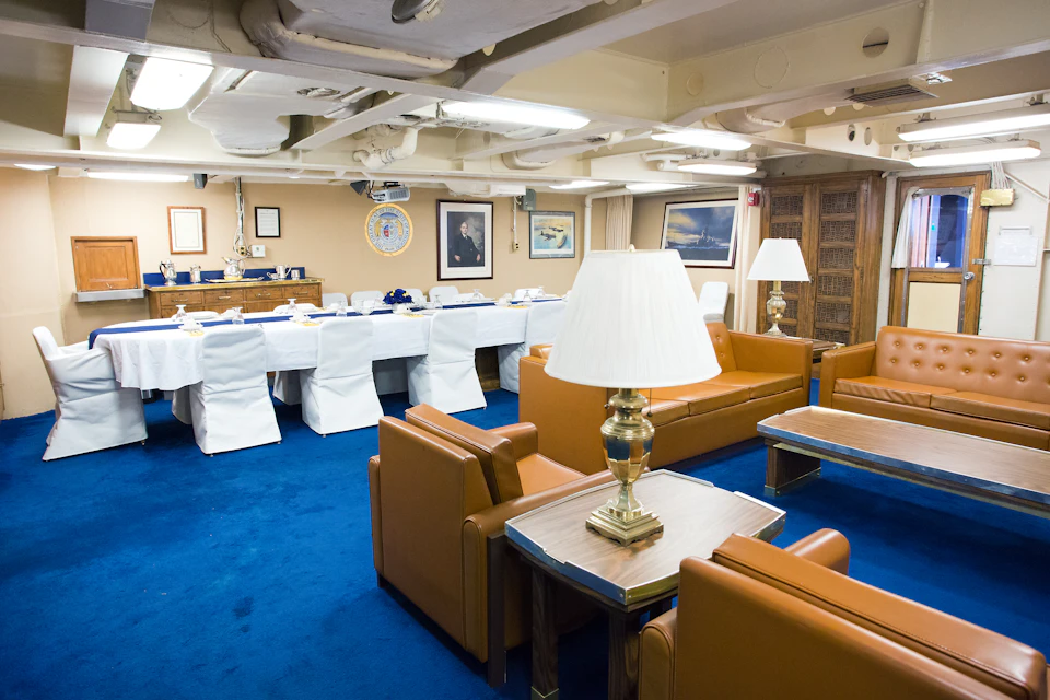 Room with blue carpet, tan leather chairs, a long table set for dining with white linens, and framed pictures on the walls.