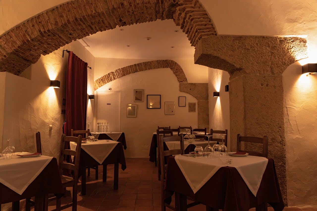 Útulný interiér restaurace s cihlovými oblouky, zarámovanými obrazy na stěnách, dřevěnými židlemi a stoly s bílými a hnědými ubrusy.