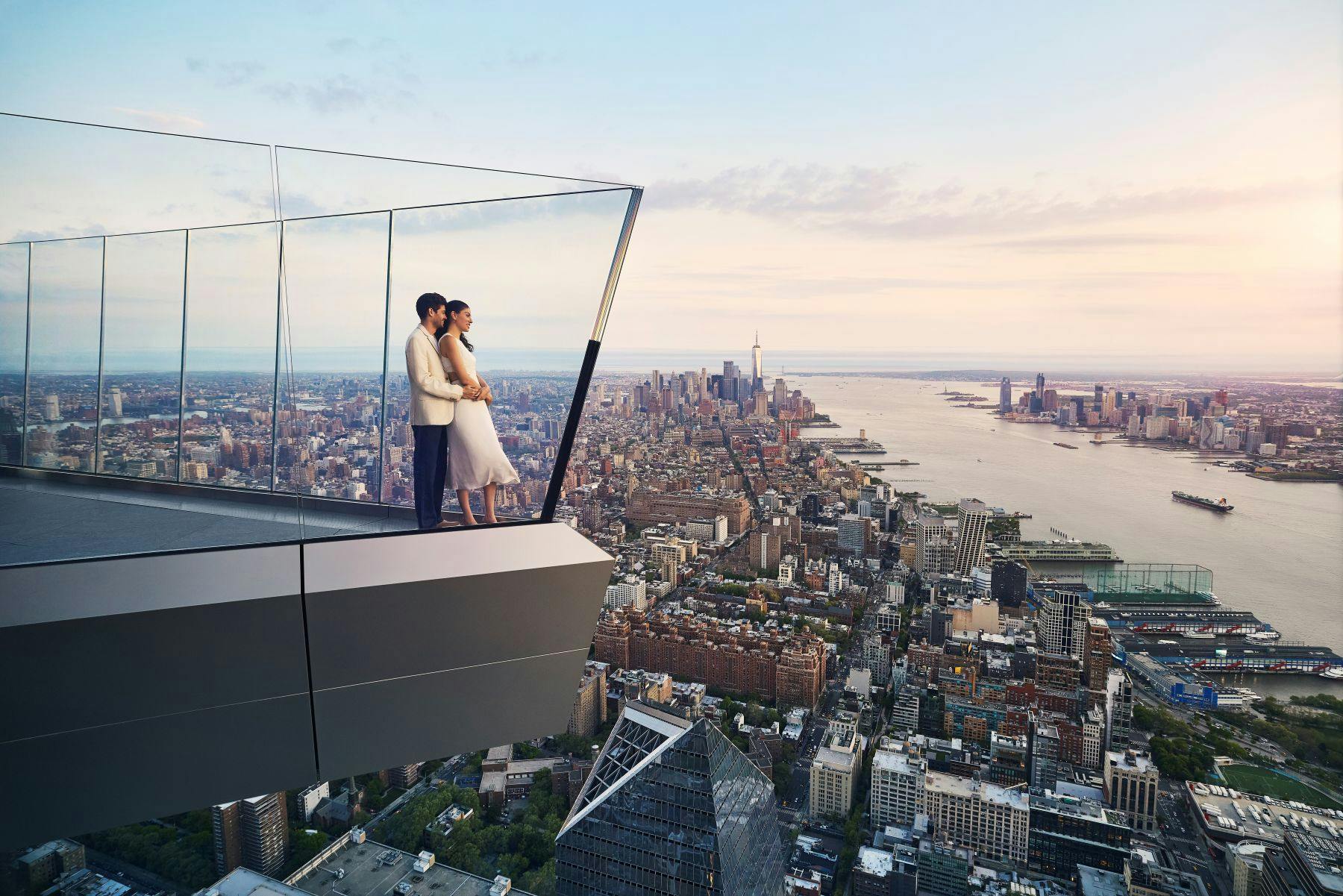 Un couple se tient sur une terrasse d'observation en verre surplombant un paysage urbain avec de grands bâtiments et une rivière au coucher du soleil.