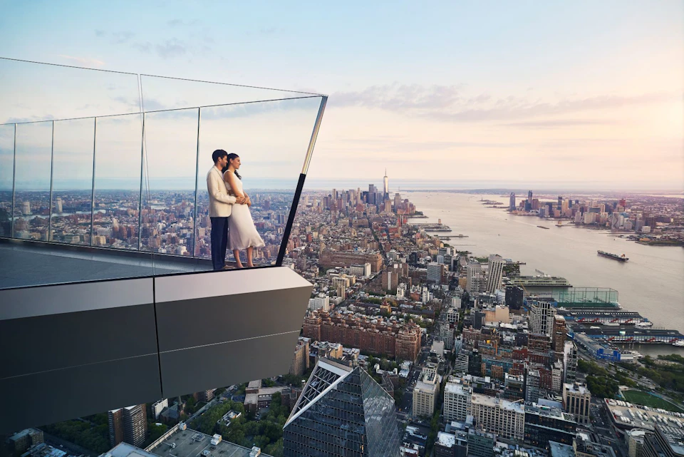 A couple stands on a glass observation deck overlooking a cityscape with tall buildings and a river at sunset.
