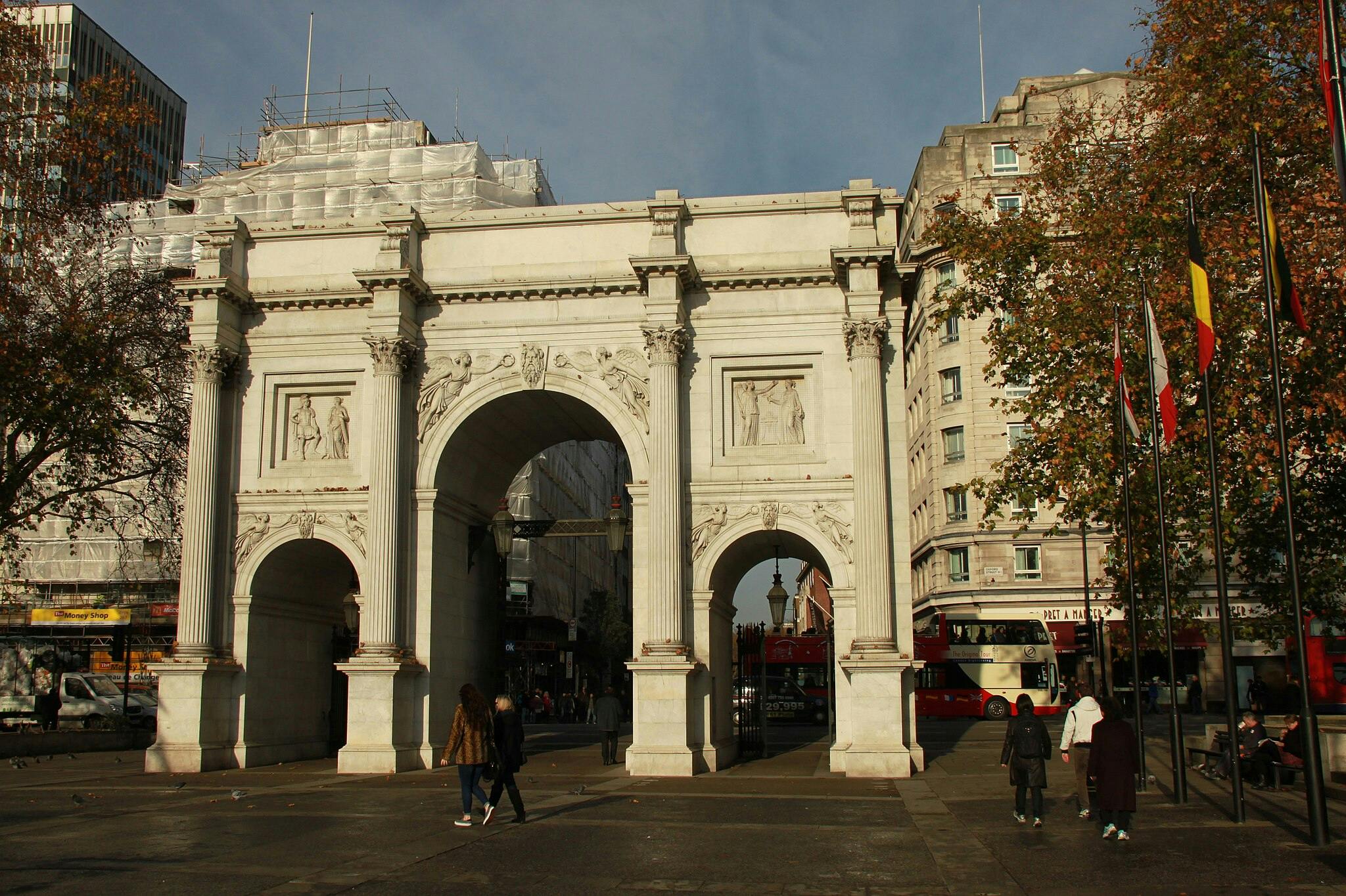 Lidé kráčející kolem velkého bílého mramorového oblouku se složitými řezbami, obklopeni budovami, stromy a stožáry.