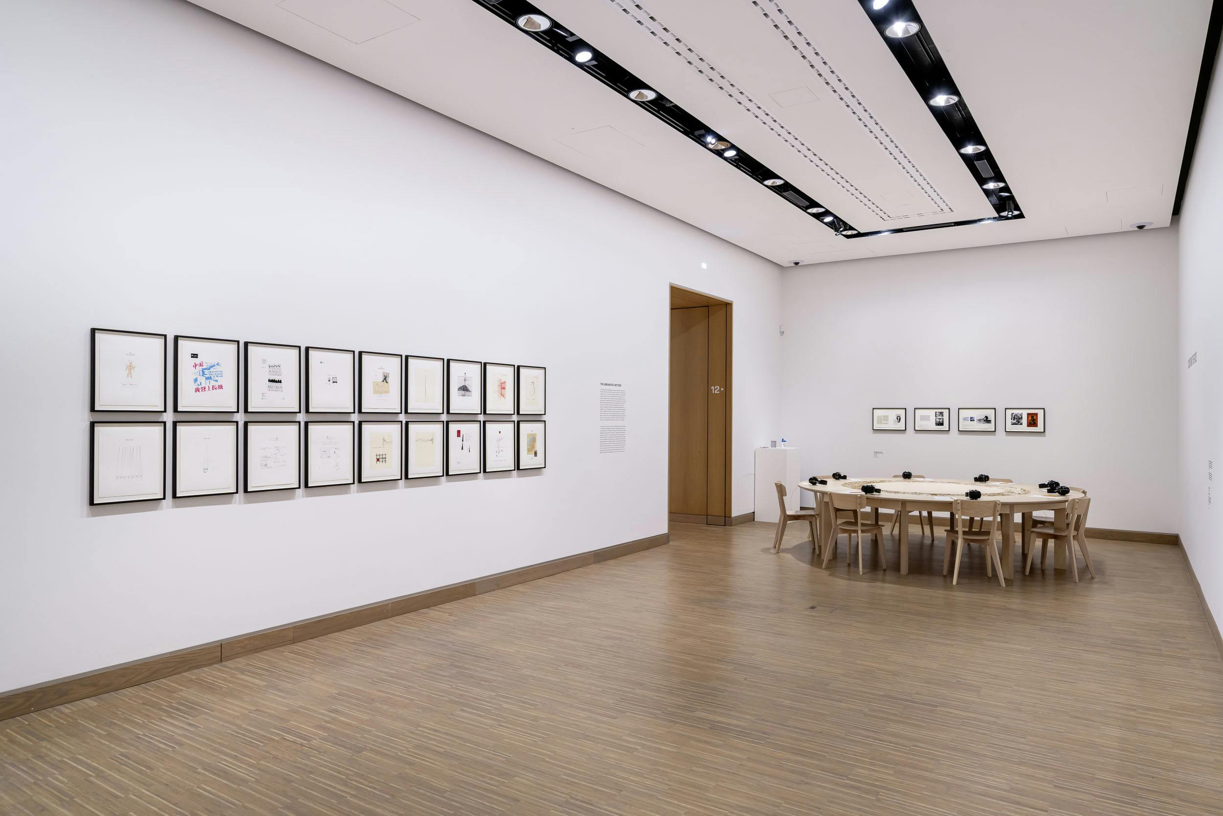 Μια σύγχρονη αίθουσα γκαλερί με κορνιζαρισμένα έργα τέχνης στους τοίχους και ένα ξύλινο τραπέζι με καρέκλες στο κέντρο.