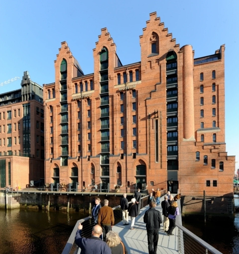 People walking on a bridge towards a large, red-brick building with Gothic-style architecture on a sunny day.