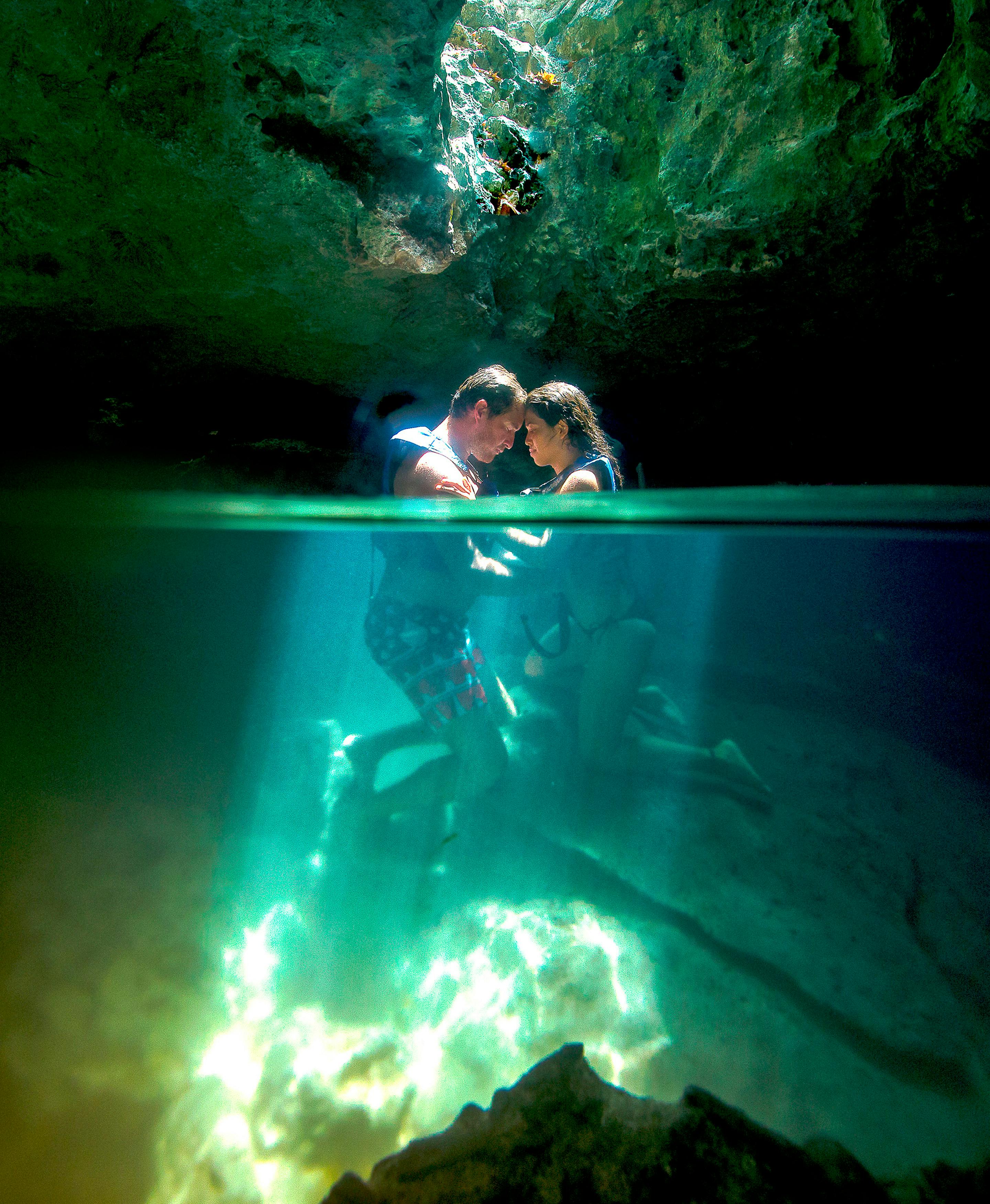 Un couple sous l'eau dans une grotte, éclairé par la lumière du soleil depuis le haut, avec des rochers et de l'eau bleue claire autour d'eux.