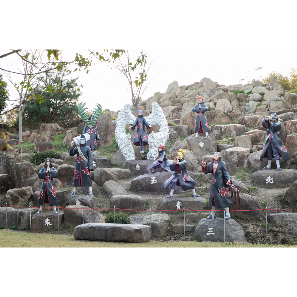 Statues of characters in black cloaks with cloud symbols, arranged on a rocky hill with some greenery and trees in the background.