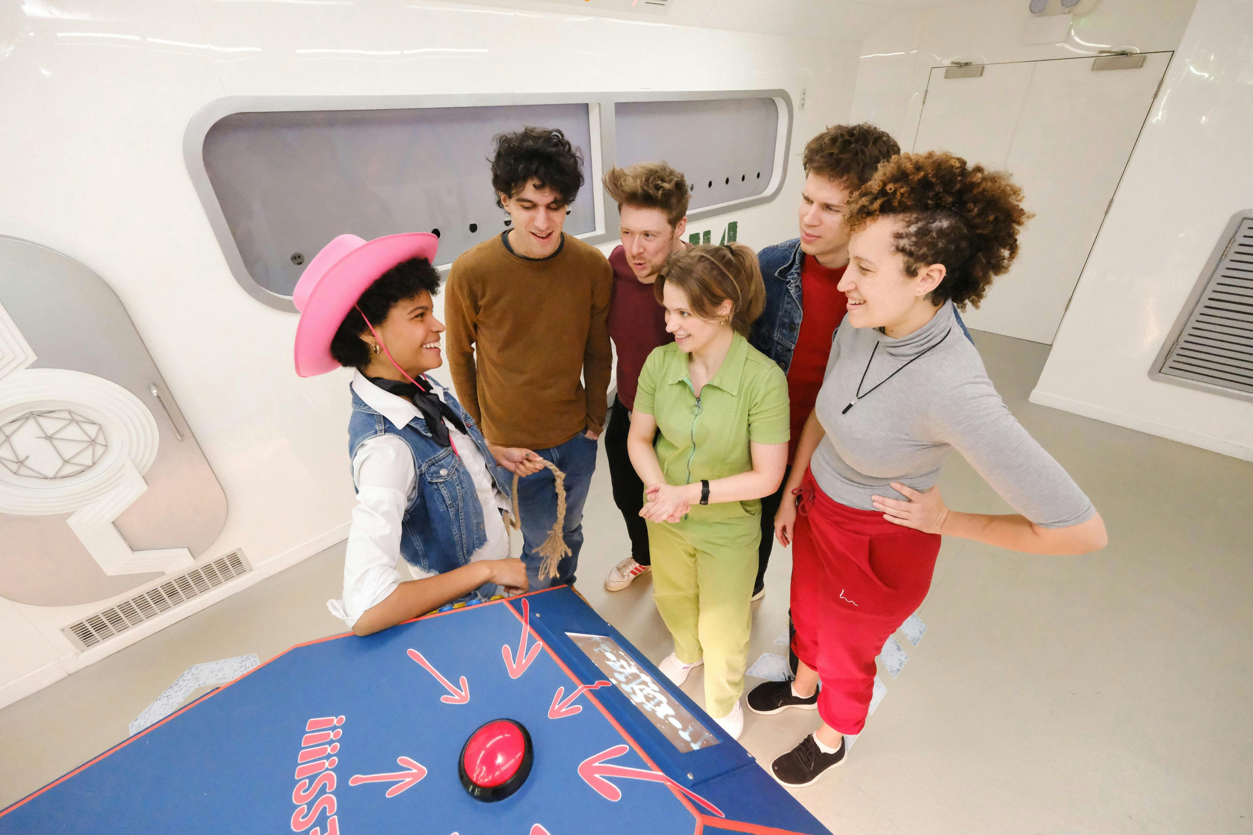 Un gruppo di cinque persone sorridenti e in piedi intorno a un tavolo con un grande bottone rosso, una persona indossa un cappello da cowboy rosa.