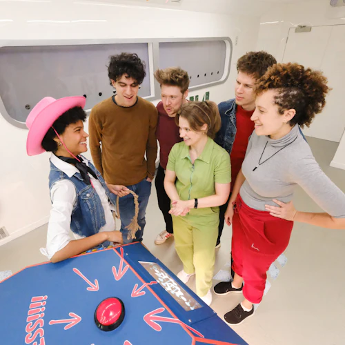 En grupp på fem personer som ler och står runt ett bord med en stor röd knapp, en person som bär en rosa cowboyhatt.