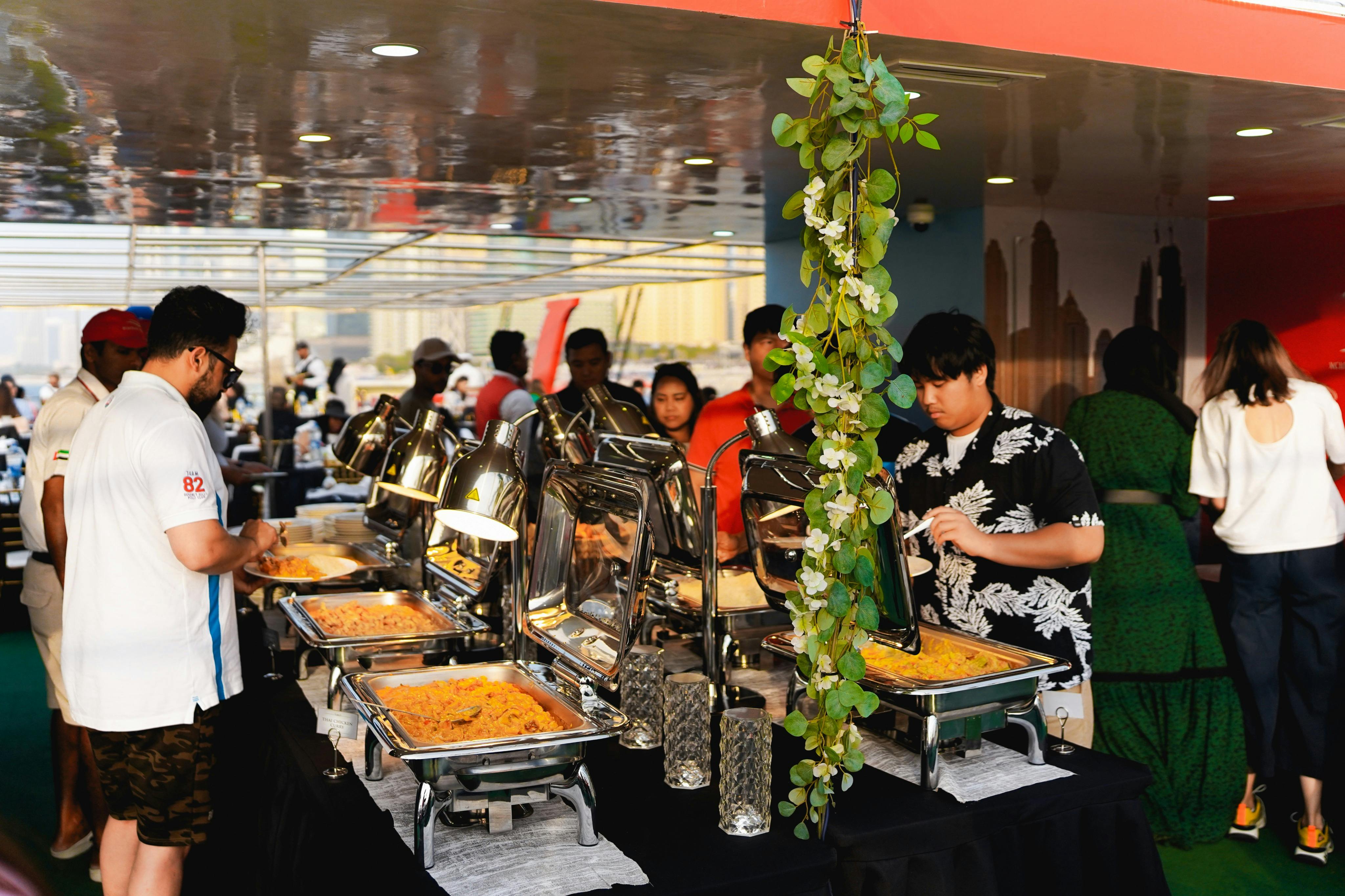 Lidé si servírují jídlo z bufetu s odřeným nádobím; nad sestavou visí zelené ozdobné listí.