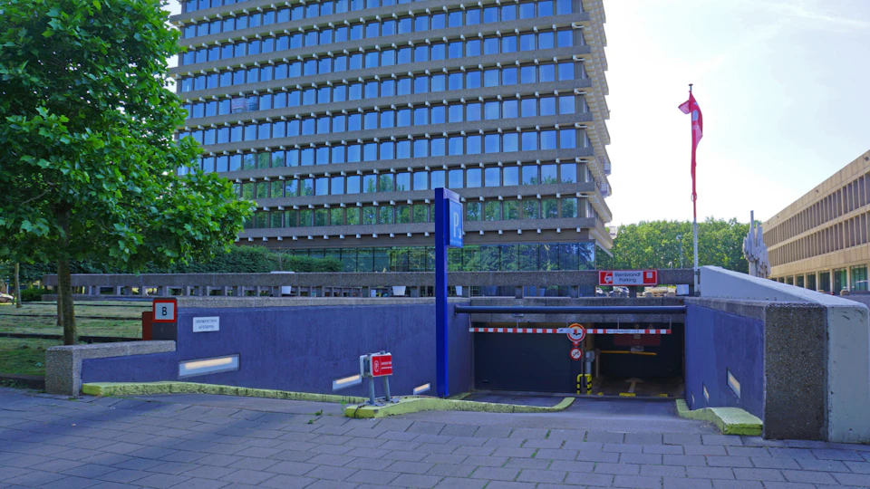 A multi-story building with reflective windows, an underground parking entrance, signage, and a few trees on the left.