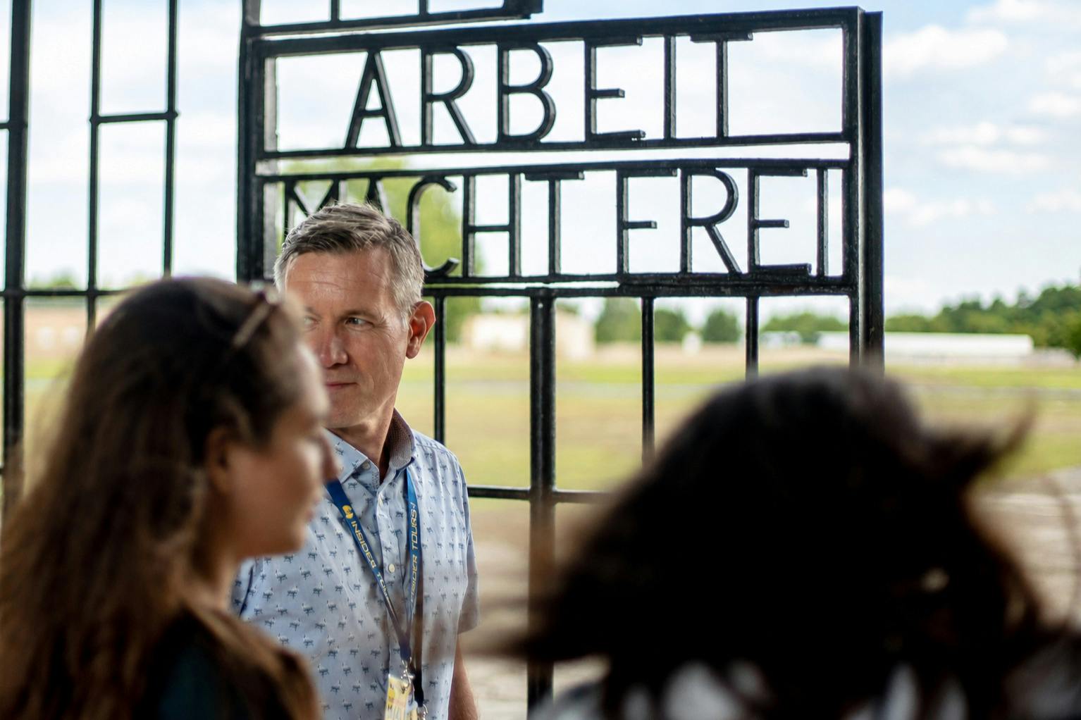 屋外で「Arbeit Macht Frei」の文字が見える歴史的な門の近くに立っている男性と他の人たち。