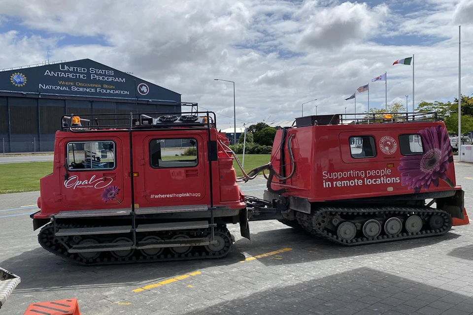 Two red tracked vehicles connected together, parked near a building with a sign for the United States Antarctic Program.