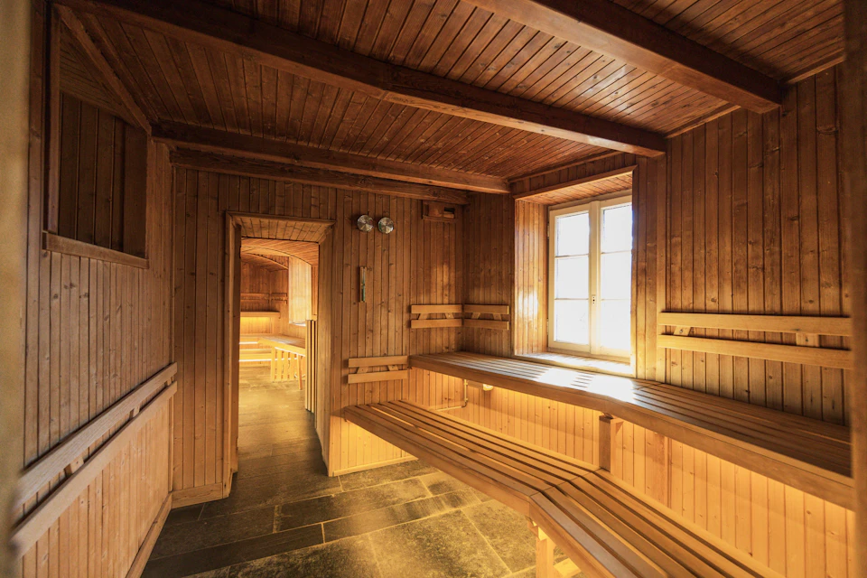 A wooden sauna with benches, a window letting in sunlight, and a doorway leading to another room.