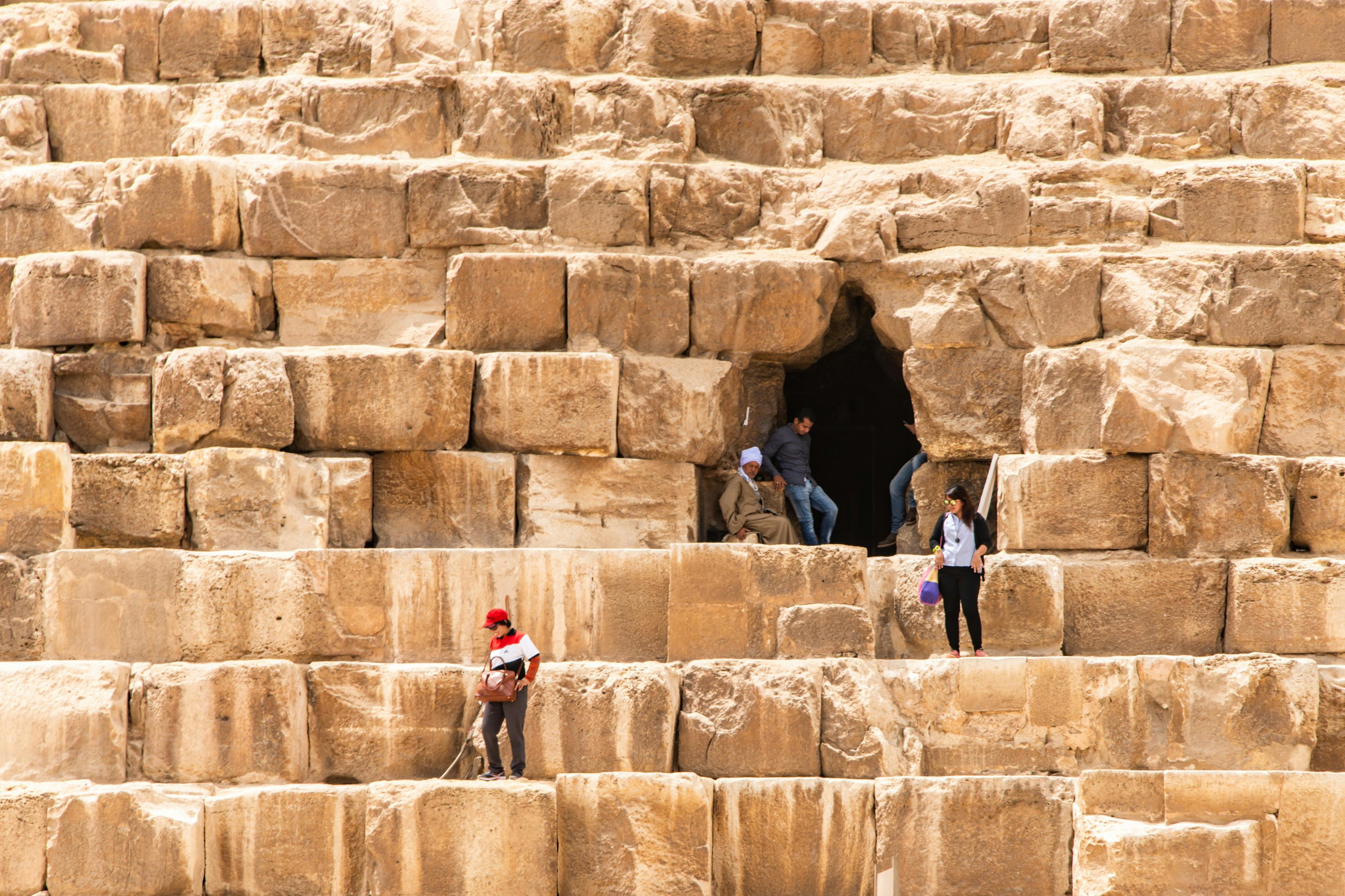 People climbing the ancient stone blocks of a pyramid, with two individuals near an entrance and one climbing with a rope.