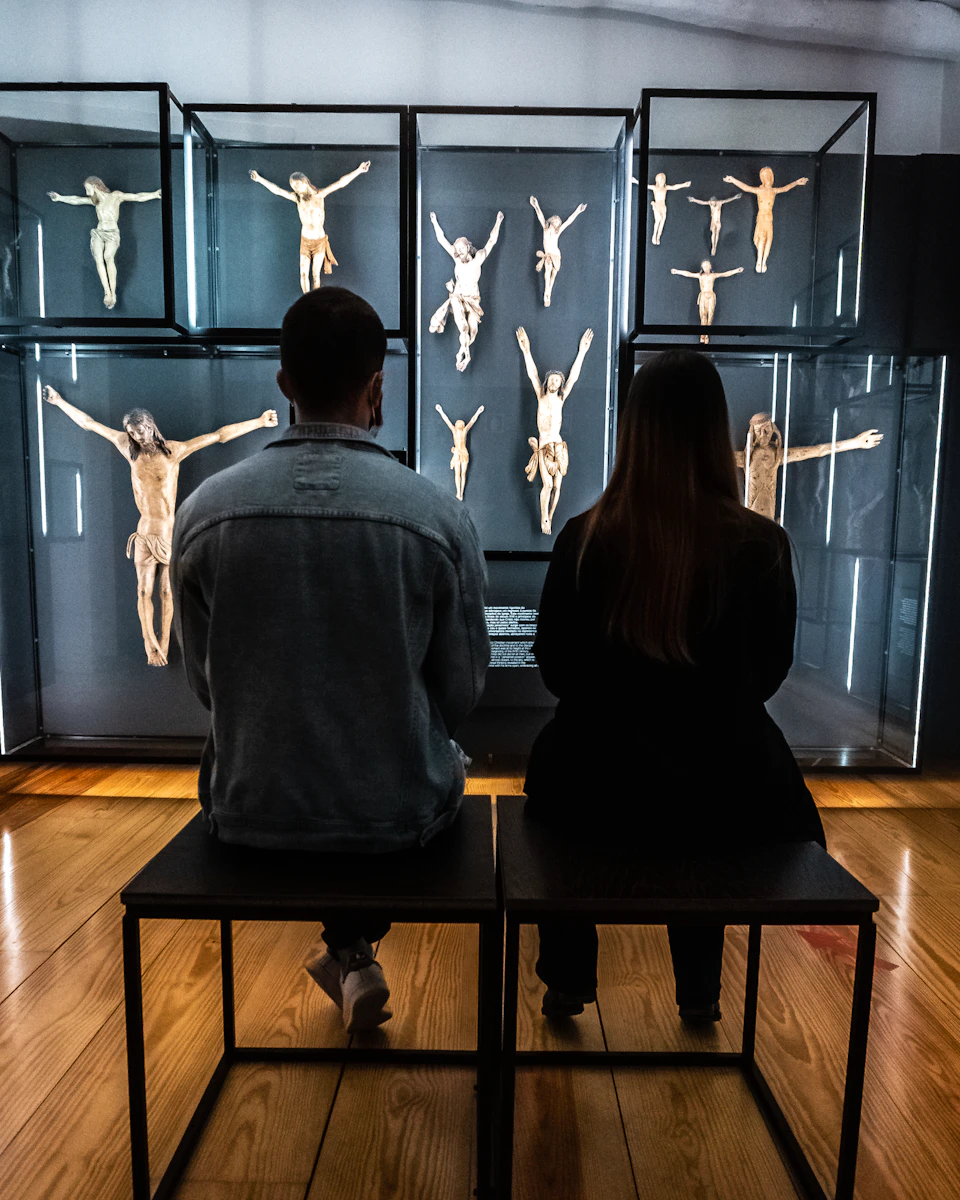 Two people seated on stools viewing a display of crucifixion sculptures in a museum.