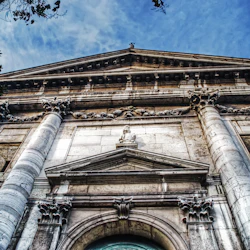 En närbild av den utsmyckade fasaden på en gammal byggnad med pelare, skulpturer och en triangulär pediment under en blå himmel.