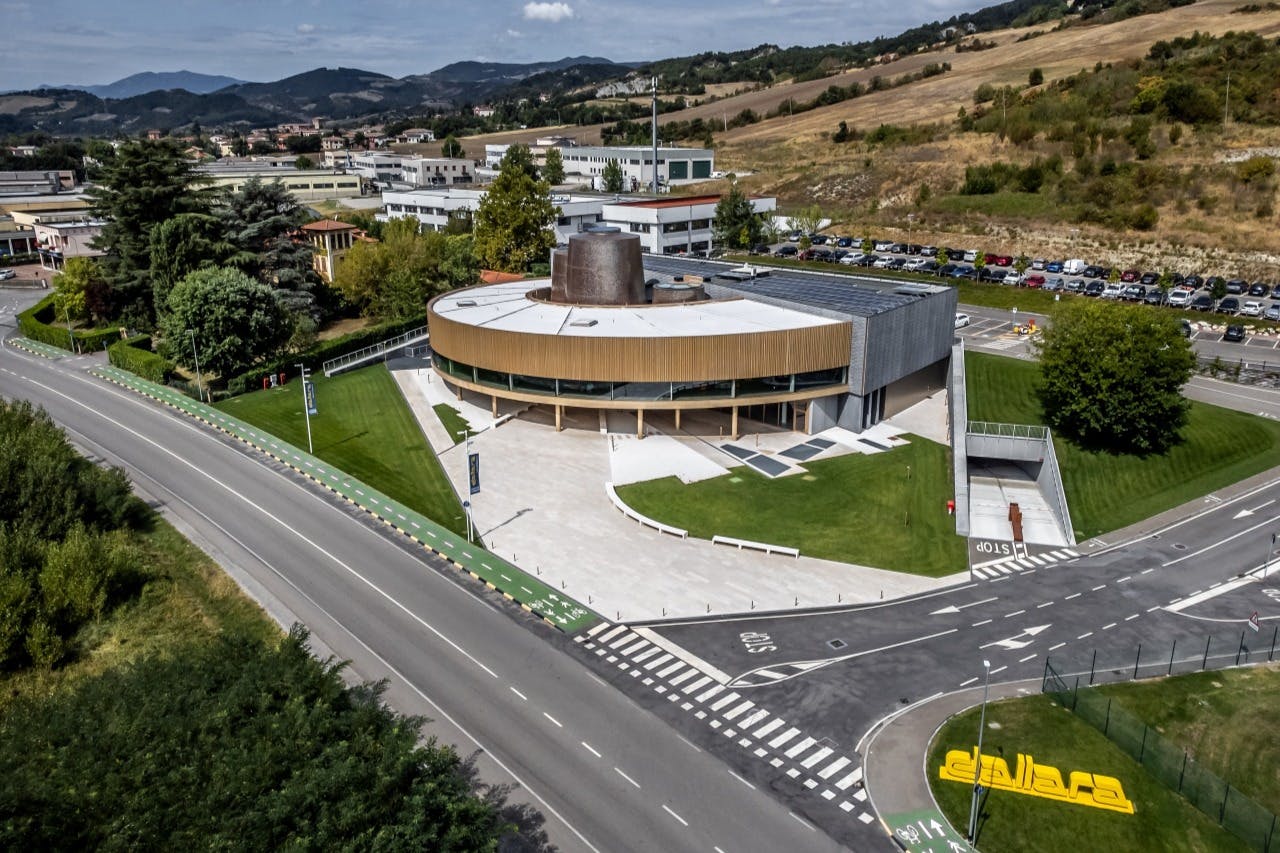 Un moderno edificio circolare con grandi finestre, circondato da strade, parcheggi e verde in una zona collinare.