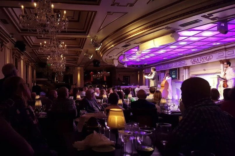 A performer on stage entertains an audience in a lavishly decorated room with chandeliers and soft, purple lighting.