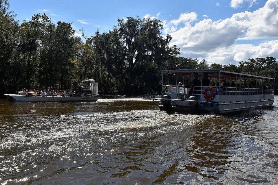 Two boats with passengers navigate a river with rippling water, surrounded by trees under a partly cloudy sky.