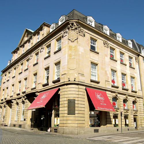 Hörnvy av en historisk beige byggnad med röda markiser och flera fönster, belägen på en kullerstensgata under en klarblå himmel.