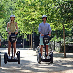 Tre personer som åker Segways i en park, med hjälmar, omgivna av gröna träd och människor som går i bakgrunden.