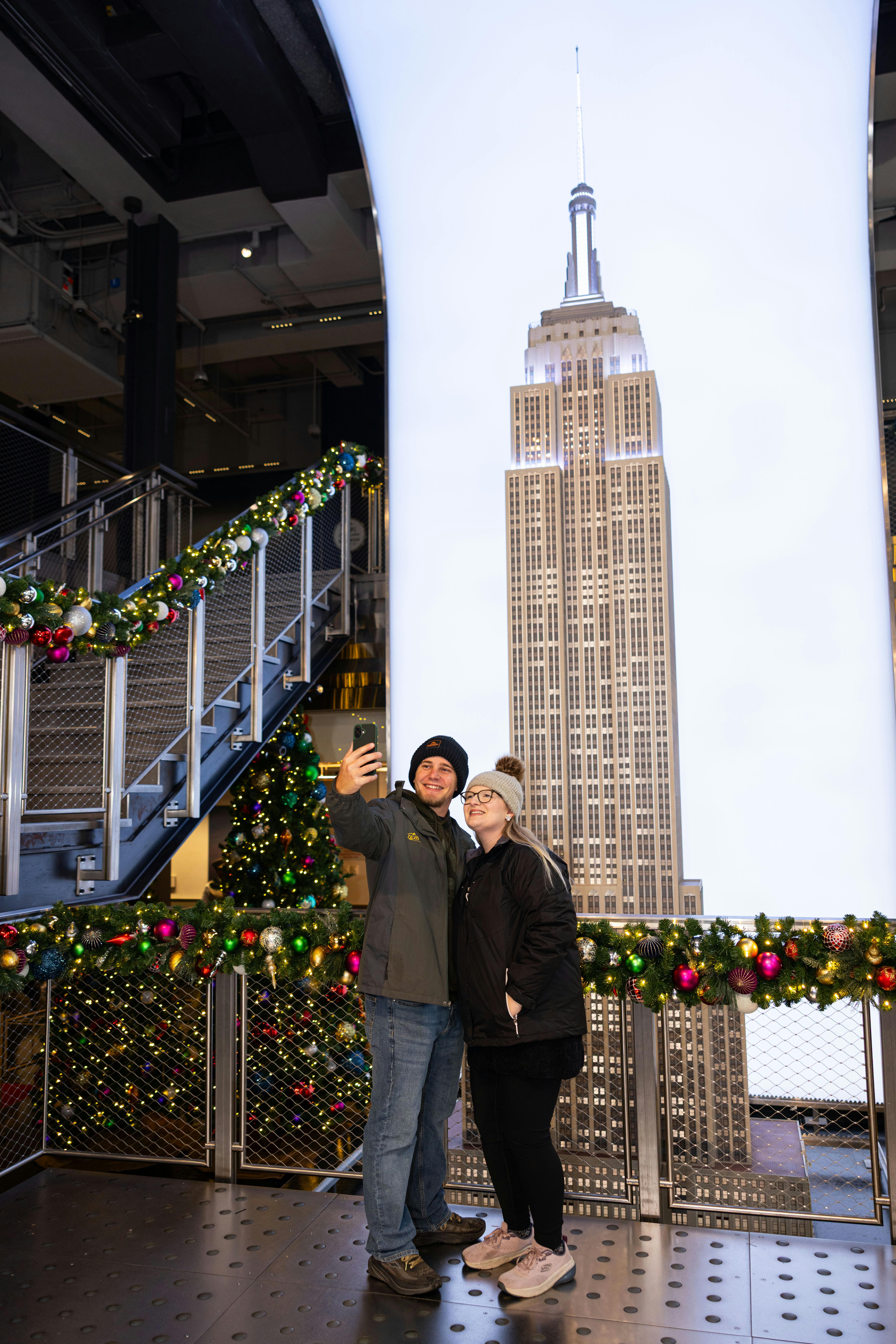 Deux personnes prennent un selfie devant une grande image de l'Empire State Building, avec des décorations festives autour d'elles.
