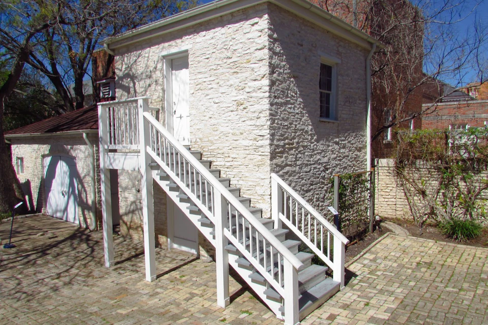 Small stone building with a white staircase leading to an upper door. Brick patio and sparse trees are visible around it.