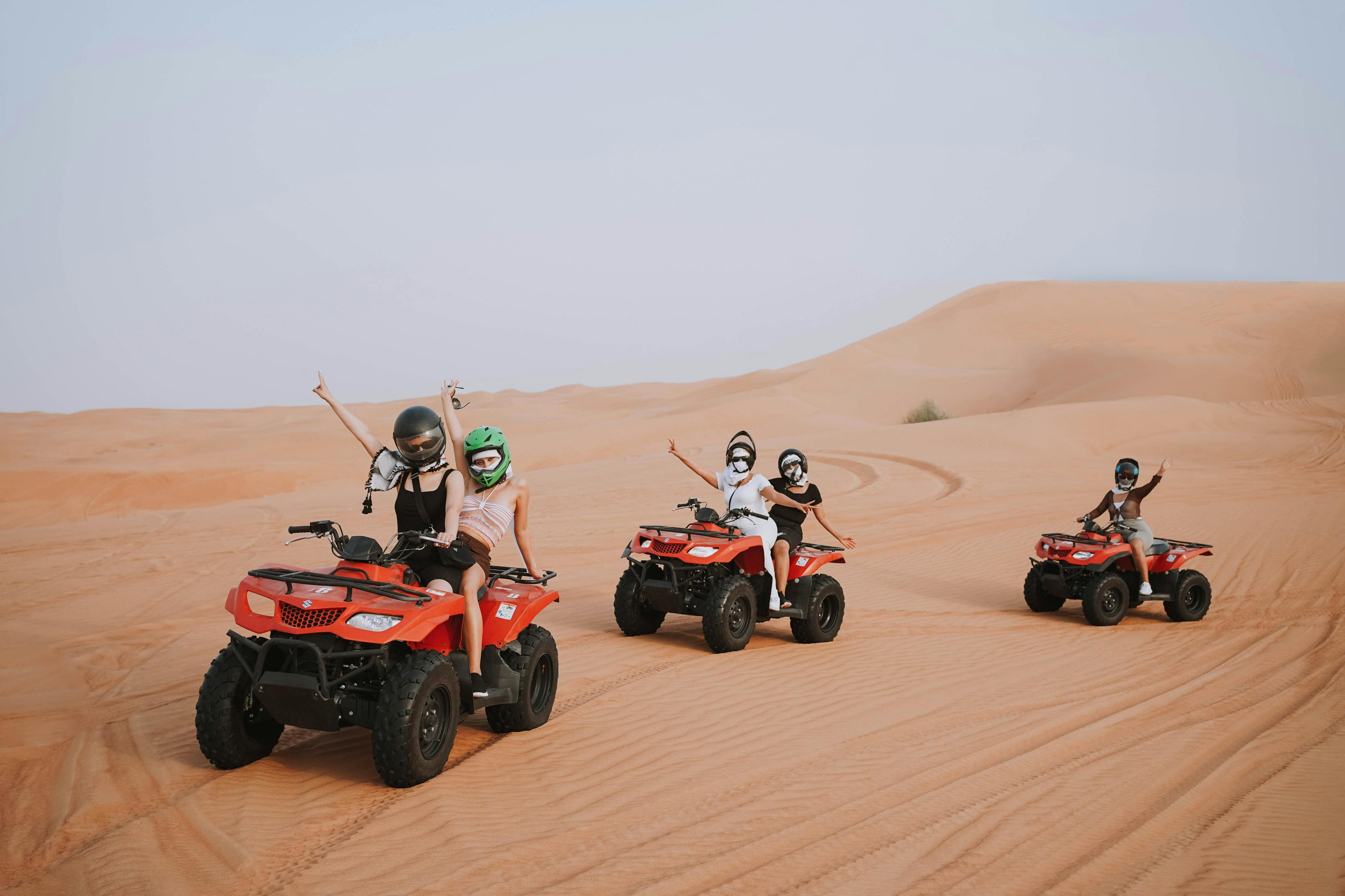 Four people riding red ATVs through desert dunes, wearing helmets and raising their arms, with sand tracks visible behind them.