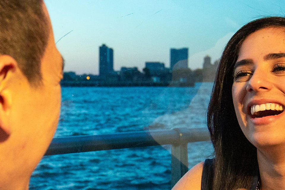A couple holds hands and smiles at a table near a window with a view of the Statue of Liberty and waterfront.