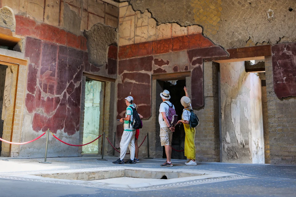 Three tourists with backpacks and hats observe ancient painted walls inside a historical site with roped-off sections.