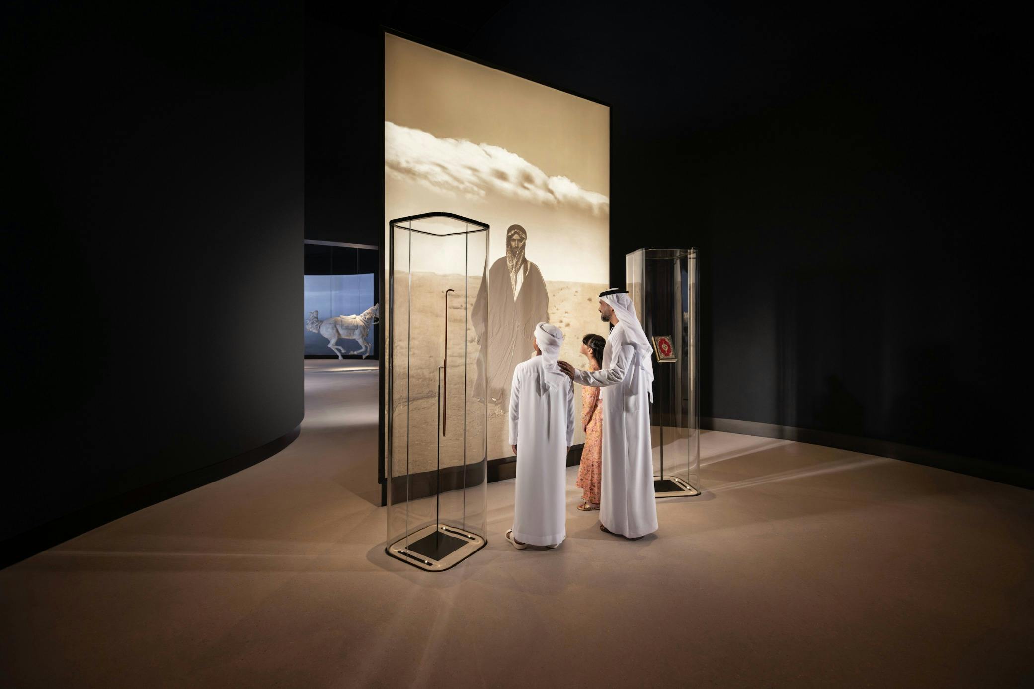 세피아 톤의 대형 초상화와 전시 케이스를 비롯하여 전통 복장을 한 세 사람이 박물관에서 전시를 관람합니다.