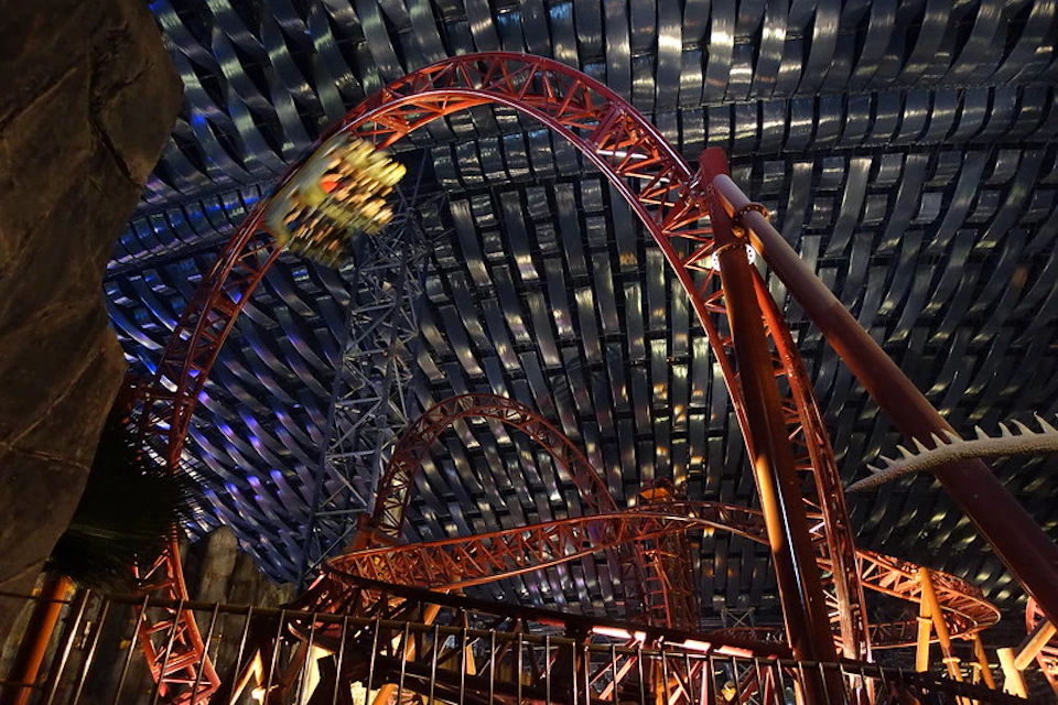 A red rollercoaster with a loop under a ribbed metal roof, featuring a blurred train in motion.