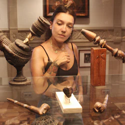 En kvinna i ett museum observerar ett skyltfodral med utsmyckade, snidade trärör och artefakter. Inramade konstverk är synliga i bakgrunden.