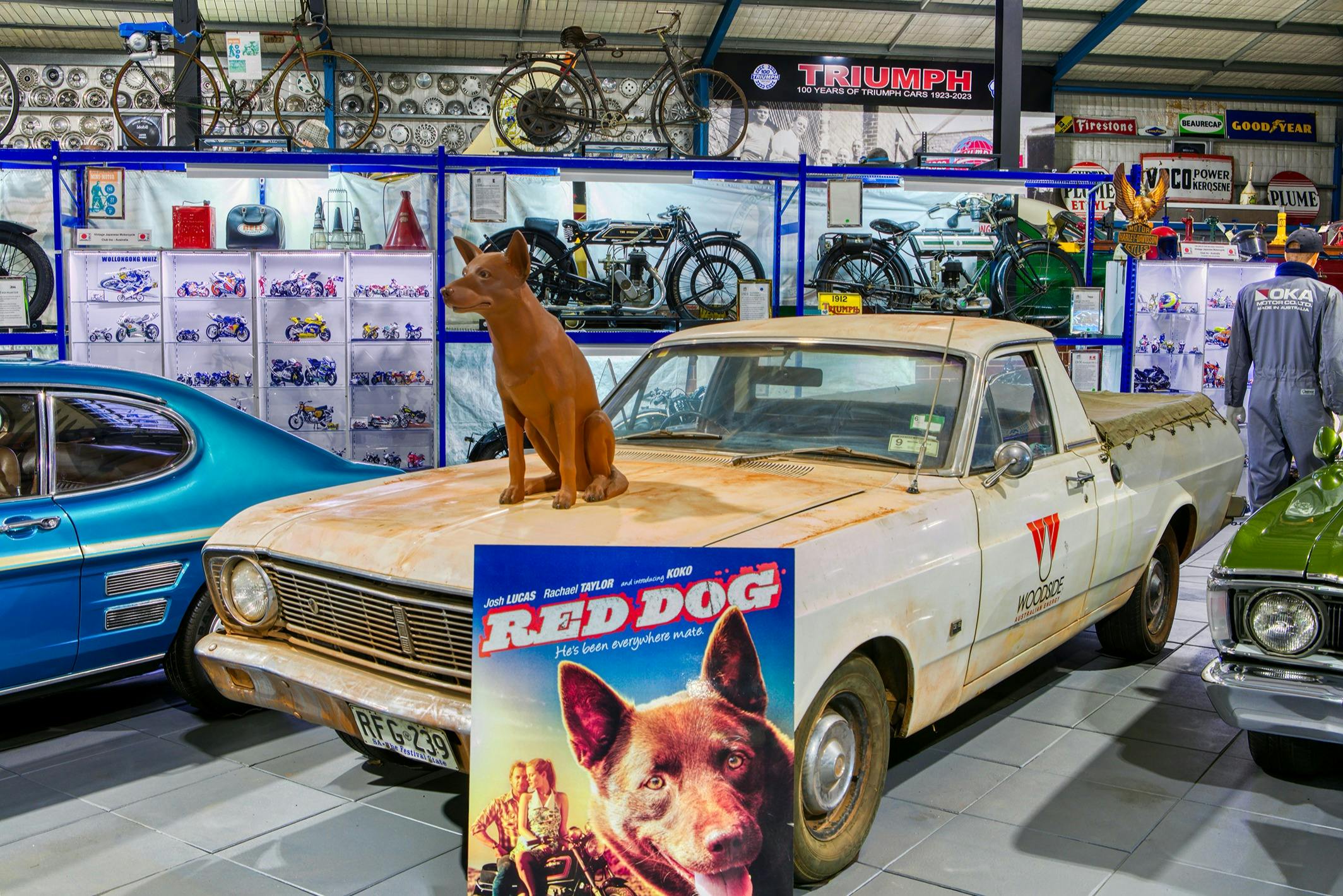 Ekspozycja muzealna ze starym białym pojazdem użytkowym z posągiem psa na masce, w otoczeniu zabytkowych rowerów i różnych przedmiotów kolekcjonerskich.