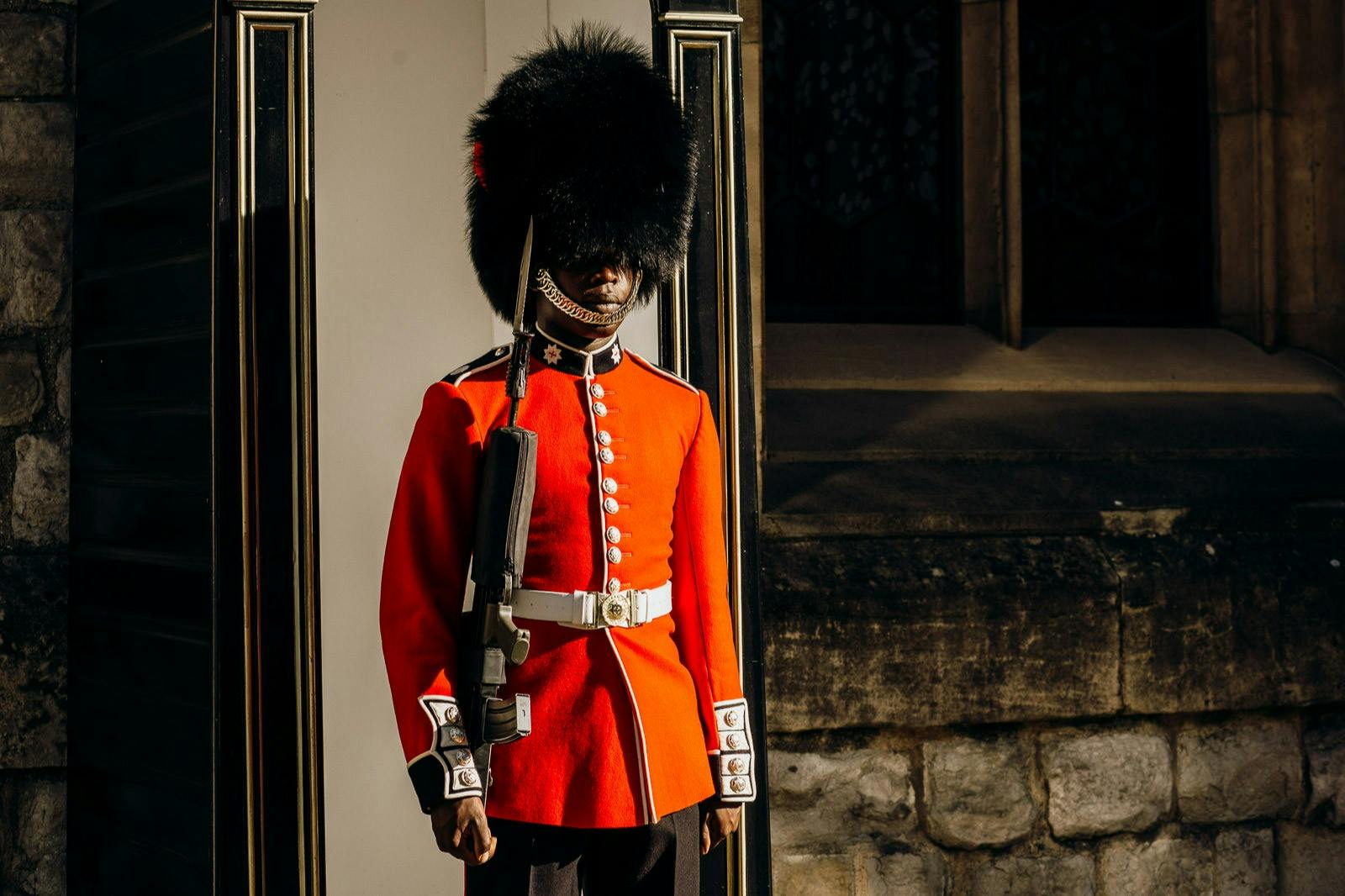 A guard in a red uniform and bearskin hat stands upright with a rifle, next to a stone wall and window.