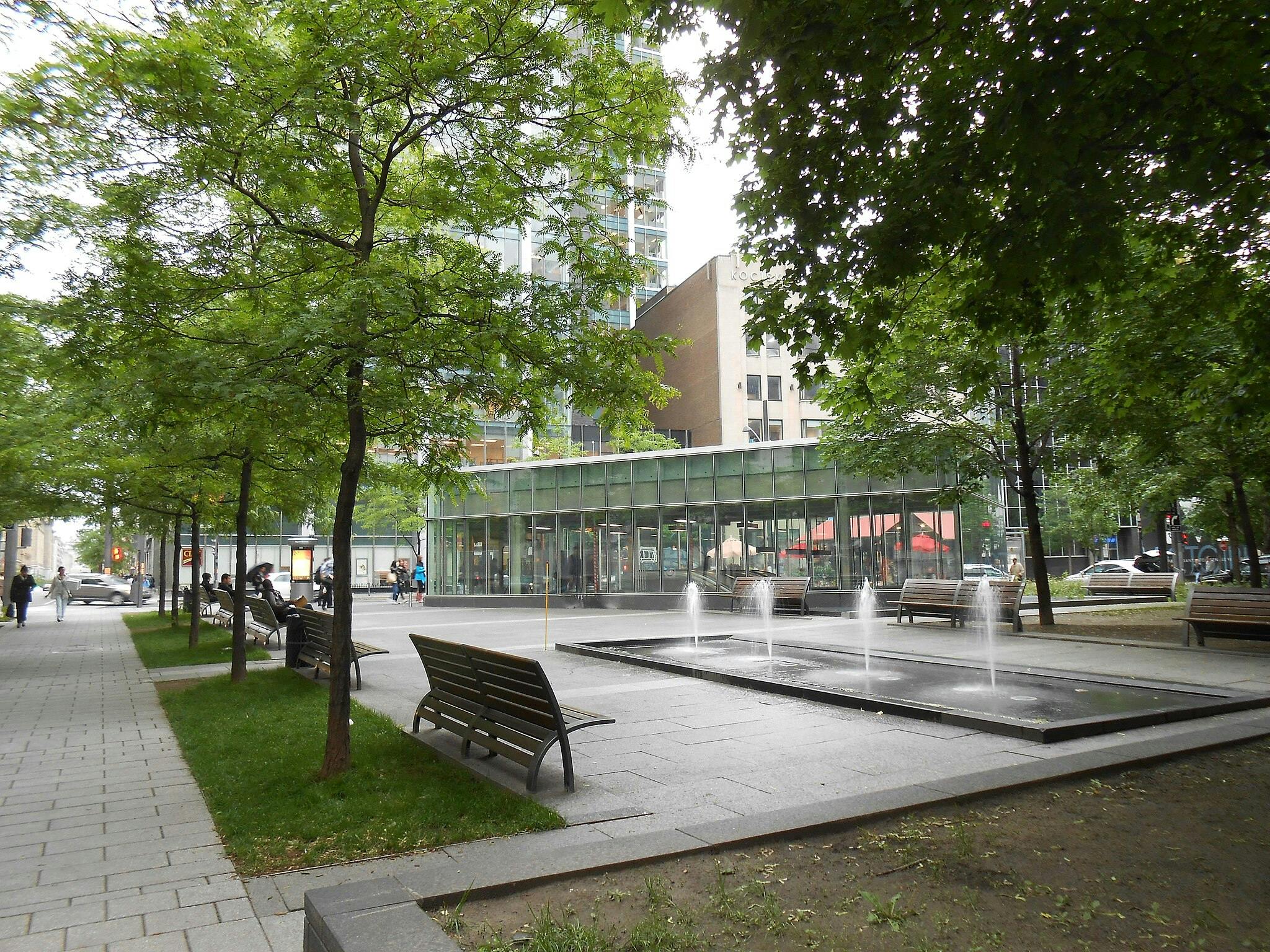Street scene with trees, benches, a small fountain, and a glass building in the background. People walking and sitting.