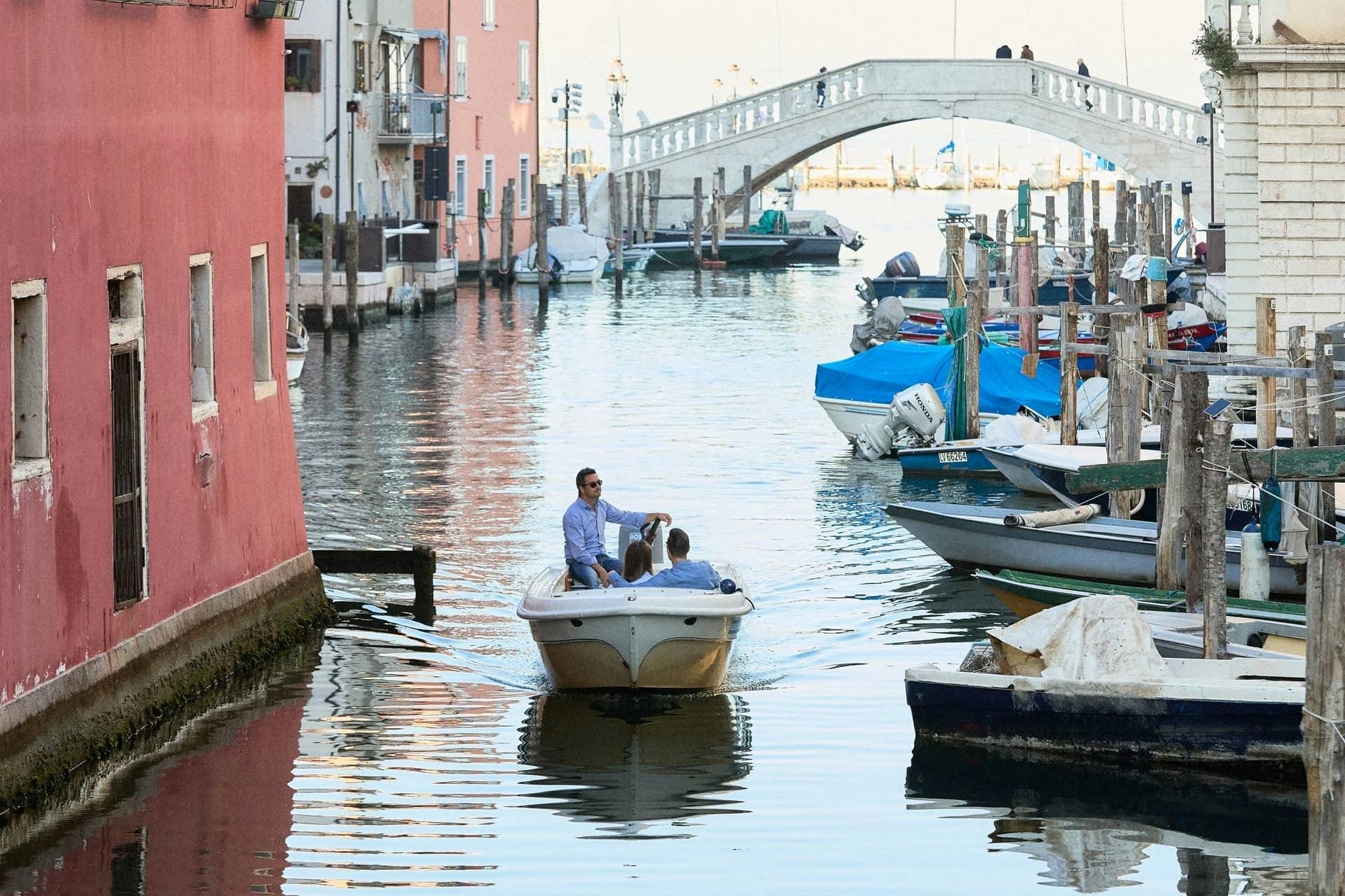 Two people are on a small motorboat navigating a canal with colorful buildings on the sides and a bridge in the background.