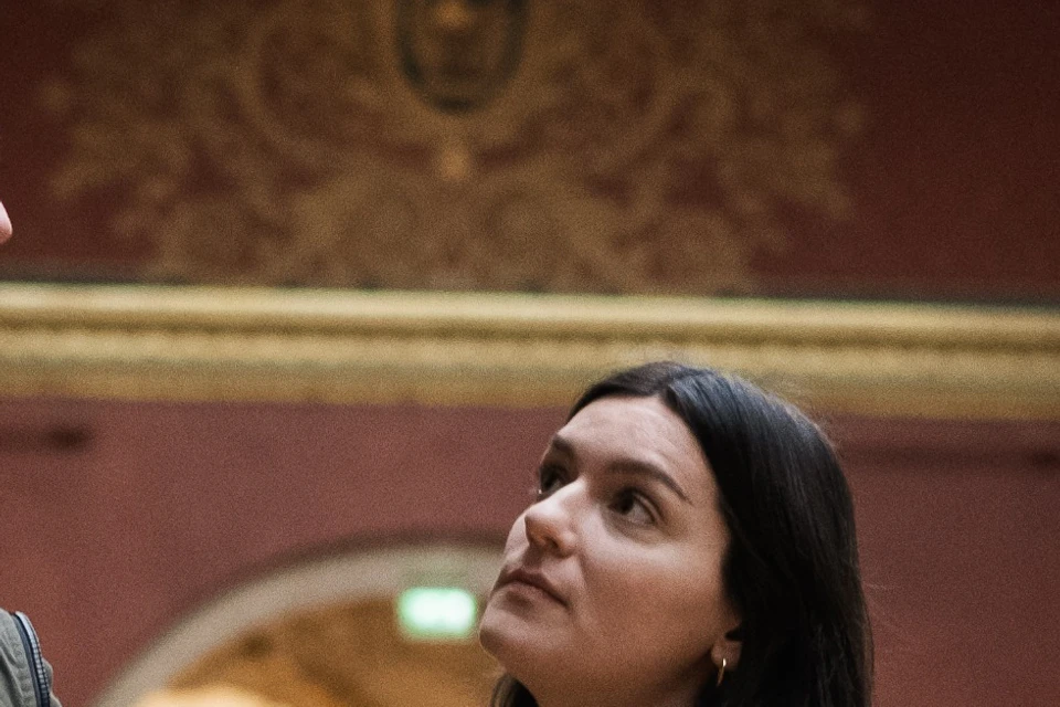 Three people are engaged in a conversation in a gallery with red walls, ornate decorations, and a large skylight.