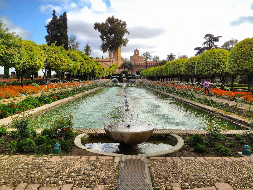 A long fountain with arched water streams, surrounded by flower beds and trimmed trees, leads towards a historic building in the background.