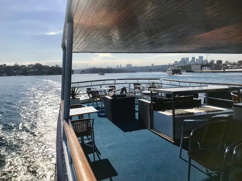 Open-air deck of a boat with chairs and tables, overlooking a city skyline and water on a sunny day.