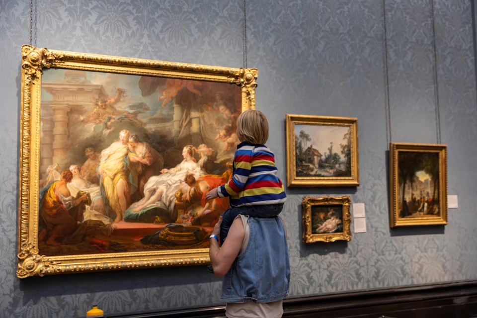 A child on an adult's shoulders viewing a large, ornate painting in an art gallery, with other framed artwork on the wall.