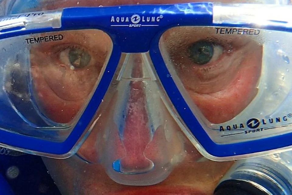 Person wearing a blue wetsuit and blue diving mask with a snorkel, partially submerged in water, looking directly at the camera.