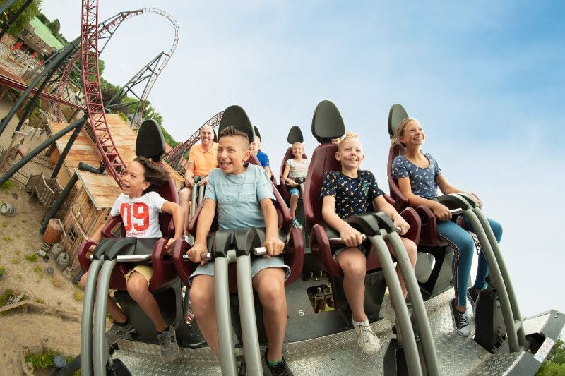 Children and adults seated on a rollercoaster, smiling and laughing against a blue sky backdrop.
