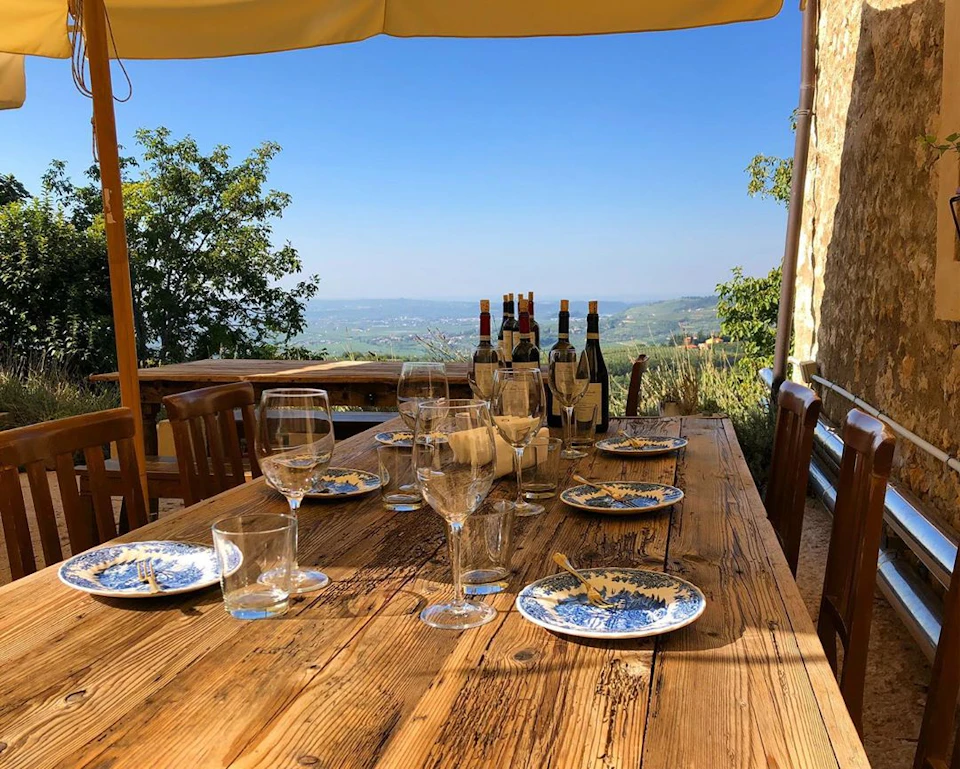 Outdoor rustic table with empty wine glasses, plates with crumbs, and several wine bottles. Scenic countryside view in the background.