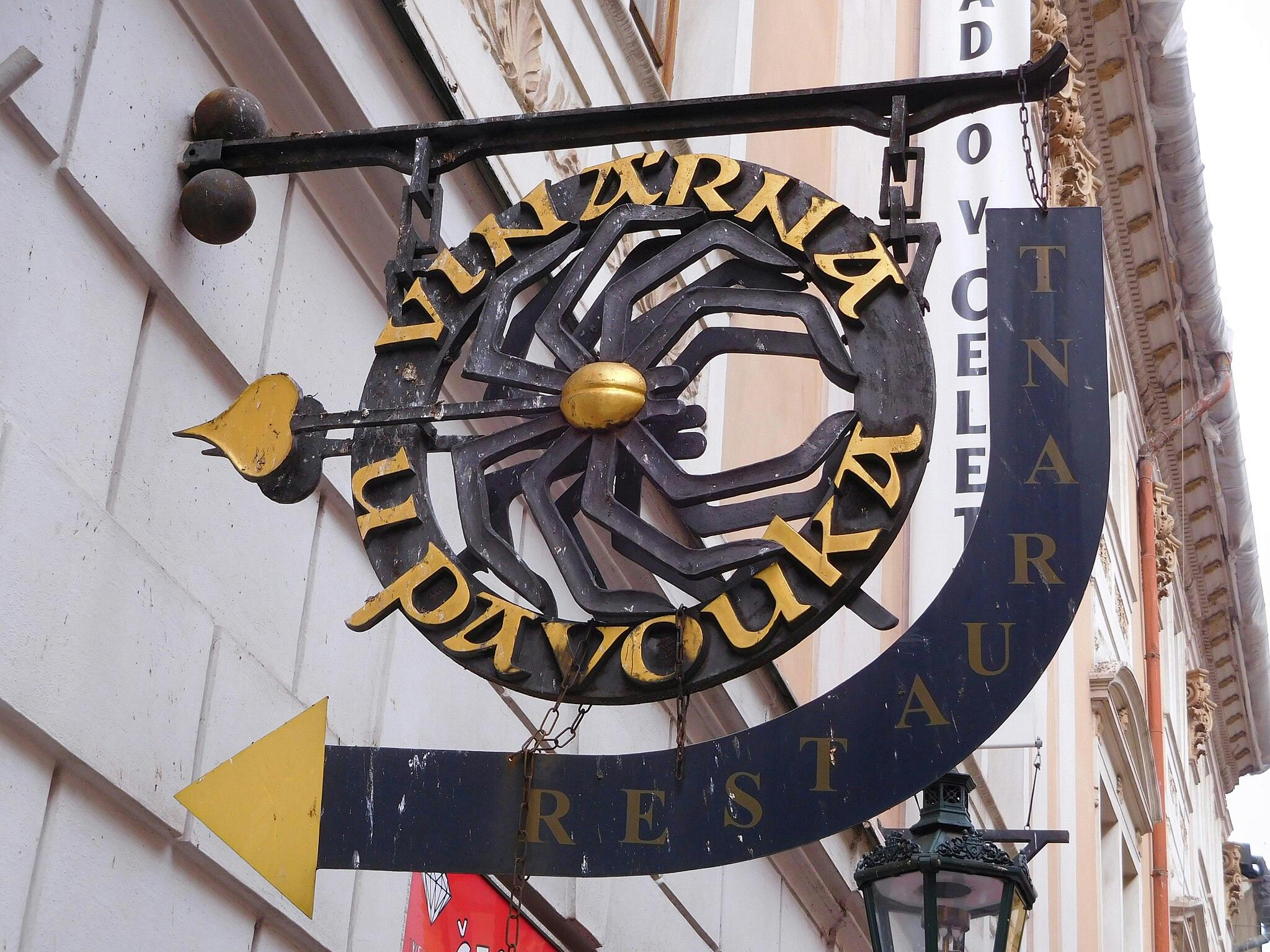 Enseigne suspendue en fer forgé indiquant "VINÁRNA U PAVOUKA" avec une flèche jaune pointant vers la gauche, montée sur la façade d'un bâtiment.