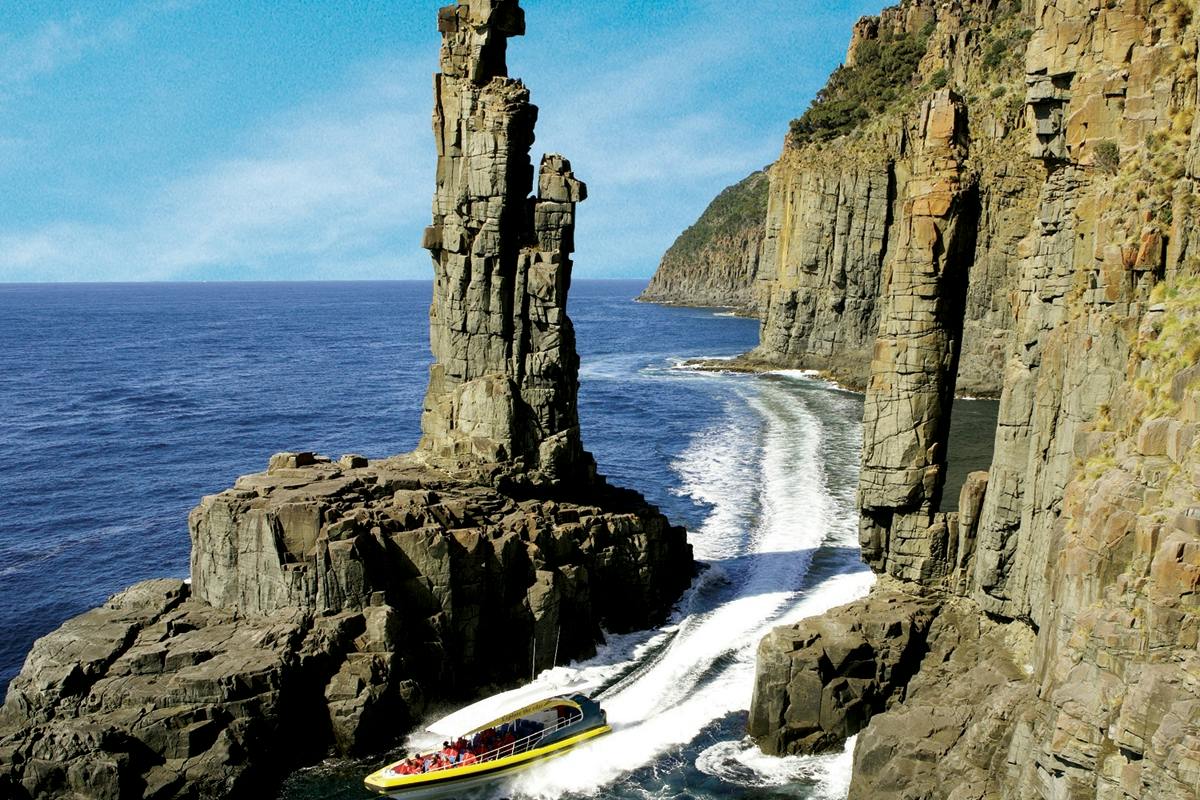 A motorboat navigates between tall coastal rock formations on a sunny day with blue ocean and cliffs in the background.