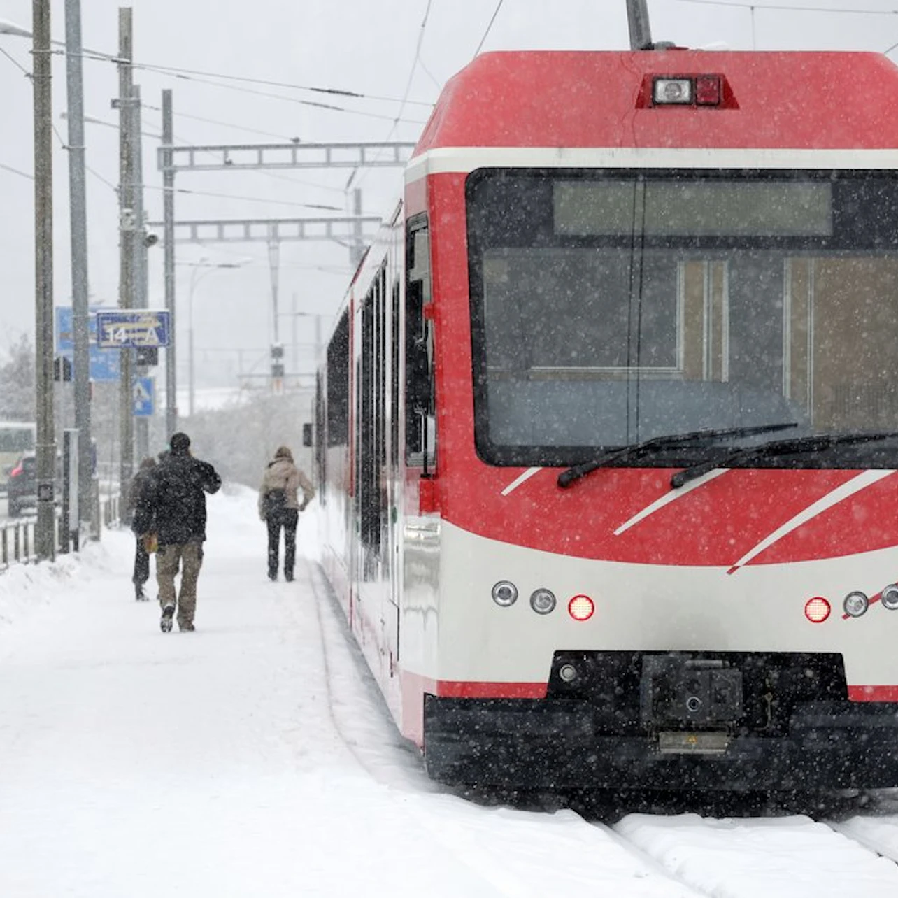 Red and white train stopped at a snowy station; three people walking through the snow along the platform.