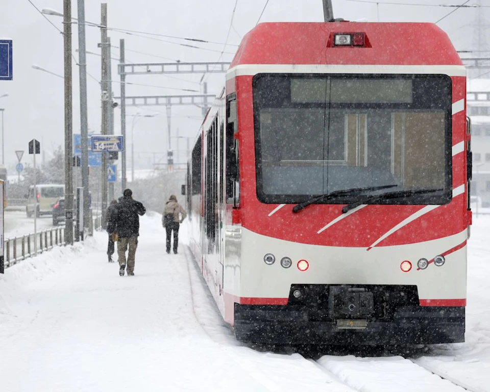 Red and white train stopped at a snowy station; three people walking through the snow along the platform.
