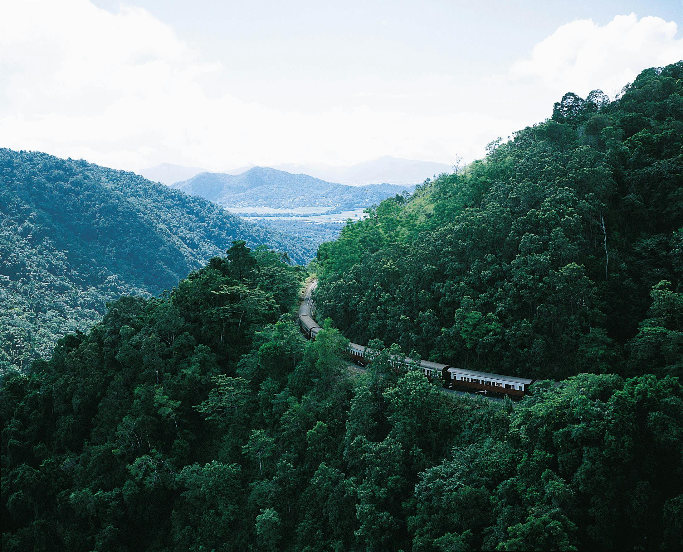 A train travels through lush, green hills with distant mountains and a cloudy sky in the background.
