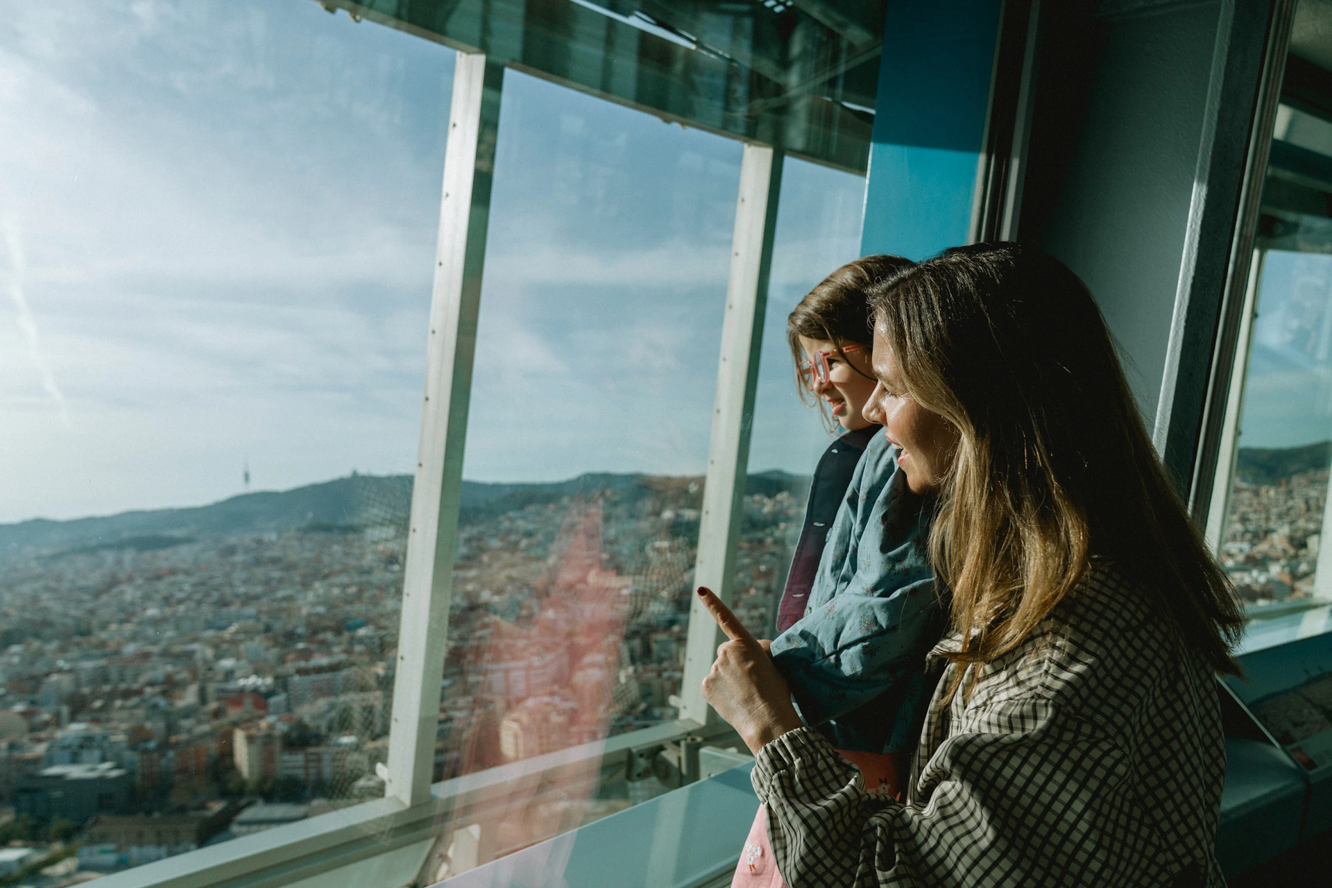 Two people are standing indoors near a large window, looking out at a panoramic view of the city beneath a bright, clear sky.